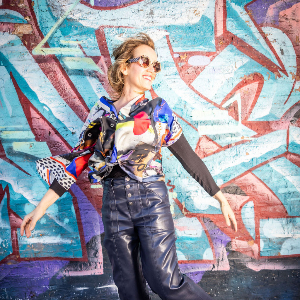 Person wearing a colorful shirt and leather pants posing in front of a graffiti wall.