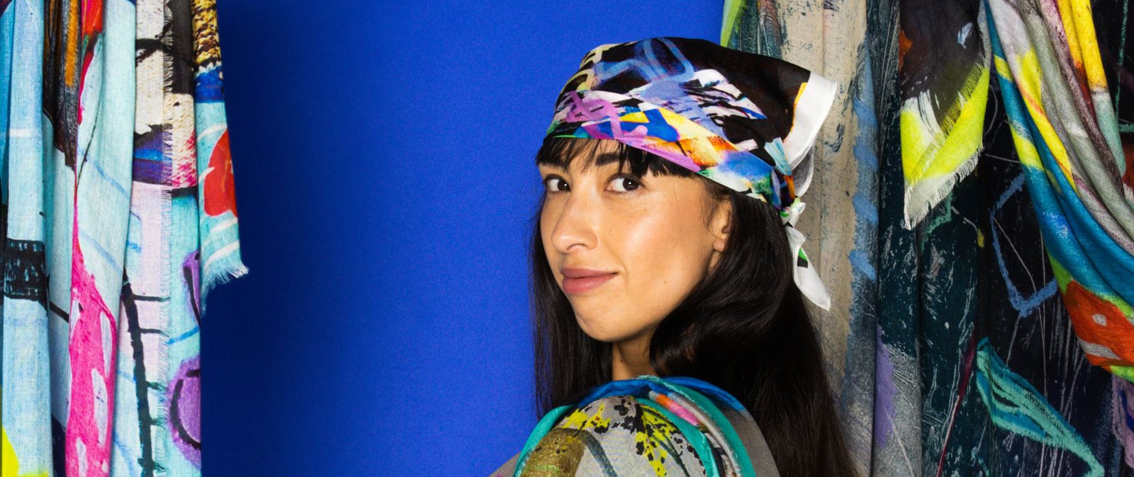 Woman wearing a colorful headscarf and scarf, standing against a vibrant graffiti wall.